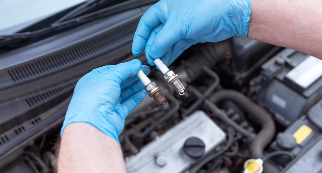 Engine Misfires and Spark Plug Issues: Mechanic holding an old spark plug in left hand and new spark plug in right hand over a car engine