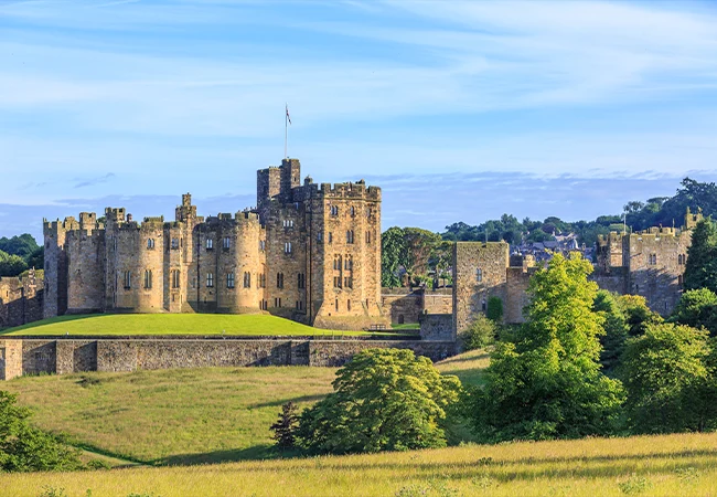 The majestic stone walls and towers of Alnwick Castle in Northumberland under a bright blue sky.