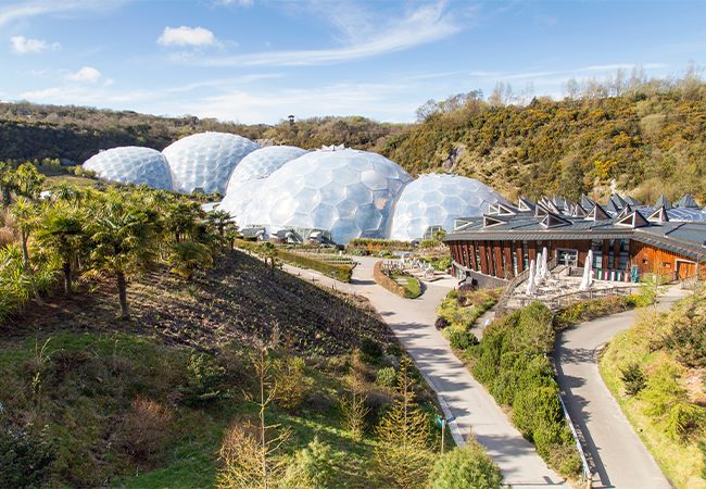 An aerial view of the massive bubble-shaped biomes at the Eden Project in Cornwall, surrounded by lush green hills.